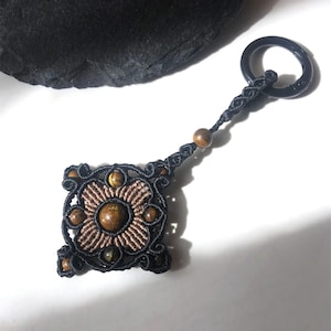 May include: A handcrafted macrame keychain featuring a square pendant with a floral design. The pendant is made with black and brown cord, and adorned with brown and gold-colored beads. The keychain has a black ring for attaching keys.