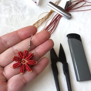 May include: Red and gold flower-shaped earring with a small red bead in the center. The earring is made with a red cord and gold thread. The earring is being held in a hand.