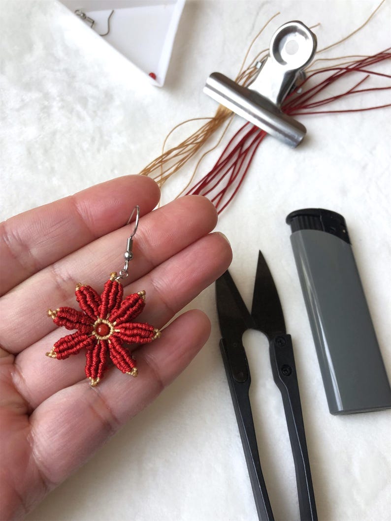 May include: Red and gold flower-shaped earring with a small red bead in the center. The earring is made with a red cord and gold thread. The earring is being held in a hand.