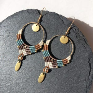 Peut inclure: Une paire de boucles d'oreilles créoles en macramé. Les boucles d'oreilles présentent des cerceaux en laiton avec un motif tissé en blanc, marron et sarcelle. De petites perles dorées et un disque doré accentuent le design. Une breloque en forme de goutte dorée est suspendue en bas.
