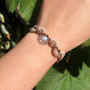 Puede incluir: Pulsera de macramé hecha a mano con una piedra lunar central rodeada de pequeñas cuentas azul claro. La pulsera tiene un diseño tejido con hilos marrones, blancos y beige, acentuado con pequeñas cuentas doradas. La pulsera está en una muñeca.