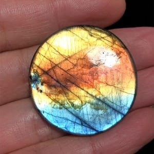 May include: A round, polished gemstone with a vibrant color gradient. The stone transitions from a warm orange and yellow to a cool blue hue. Fine lines and patterns are visible across the surface, creating a unique, natural design. The gemstone is held in a hand.