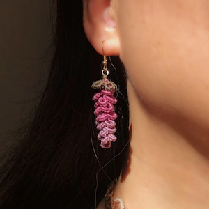 Peut inclure: Boucle d'oreille unique faite à la main avec un motif floral. La boucle d'oreille est fabriquée avec du fil rose et marron, suspendue à un crochet doré. Le travail du fil détaillé donne un aspect texturé et superposé, semblable à une fleur.