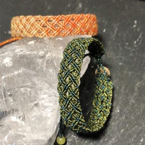 K&ouml;nnte beinhalten: Zwei gewebte Makramee-Armb&auml;nder mit komplizierten Gittermustern. Ein Armband ist aus schimmerndem orangefarbenem und cremefarbenem Garn gefertigt, w&auml;hrend das andere aus dunkelgr&uuml;nem und olivgr&uuml;nem Garn besteht und einen kleinen runden Verschluss hat.