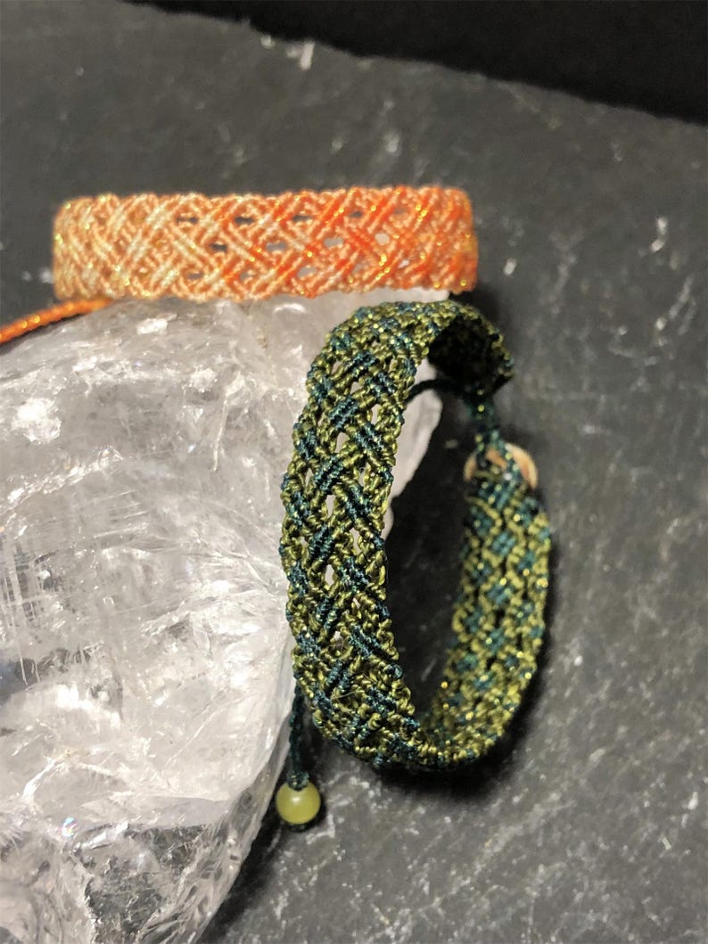 K&ouml;nnte beinhalten: Zwei gewebte Makramee-Armb&auml;nder mit komplizierten Gittermustern. Ein Armband ist aus schimmerndem orangefarbenem und cremefarbenem Garn gefertigt, w&auml;hrend das andere aus dunkelgr&uuml;nem und olivgr&uuml;nem Garn besteht und einen kleinen runden Verschluss hat.