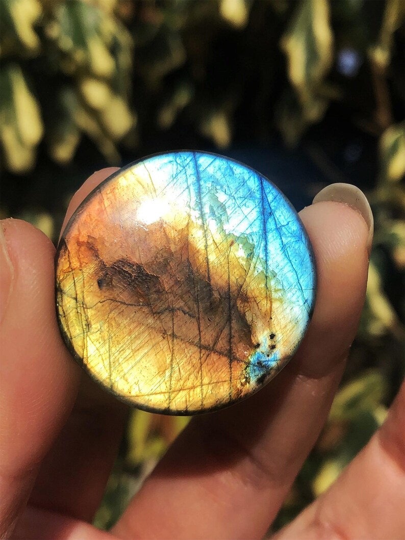 May include: A round, polished labradorite stone with a vibrant display of colors. The stone features a mix of blue, gold, and brown hues, with a shimmering, iridescent effect. The gemstone is held between fingers, showcasing its natural beauty.