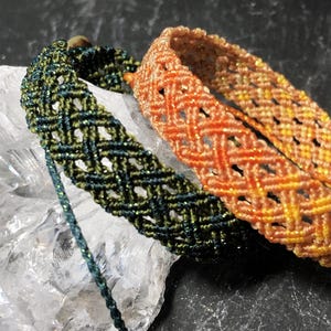 K&ouml;nnte beinhalten: Zwei Makramee-Armb&auml;nder mit einem rautenf&ouml;rmigen Gittermuster. Ein Armband ist dunkelgr&uuml;n mit dezentem metallischem Schimmer, das andere ist ein Farbverlauf aus Orange und Gelb mit Metallic-F&auml;den. Beide haben verstellbare Kordelenden.