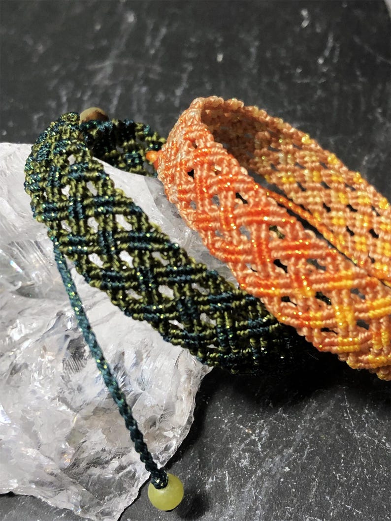 K&ouml;nnte beinhalten: Zwei Makramee-Armb&auml;nder mit einem rautenf&ouml;rmigen Gittermuster. Ein Armband ist dunkelgr&uuml;n mit dezentem metallischem Schimmer, das andere ist ein Farbverlauf aus Orange und Gelb mit Metallic-F&auml;den. Beide haben verstellbare Kordelenden.