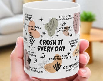 May include: White ceramic mug with the text "CRUSH IT EVERY DAY" in black, surrounded by motivational phrases and decorative elements in black, peach, and green. The mug is held in a person's hand.