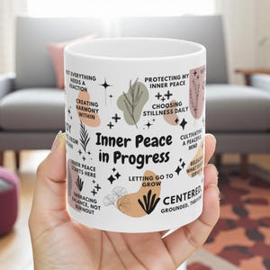 May include: White ceramic mug with the text "Inner Peace in Progress" and inspirational phrases. The mug features botanical illustrations and geometric shapes in neutral colors. The mug is held in a hand.