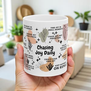 May include: White ceramic mug with the text "Chasing Joy Daily" and inspirational phrases. The mug features a design with botanical elements and geometric shapes in neutral colors. The mug is held in a person's hand.