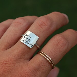 K&ouml;nnte beinhalten: Ein silberner Ring mit einem quadratischen Band, auf dem "OWN YOUR JOURNEY" steht.