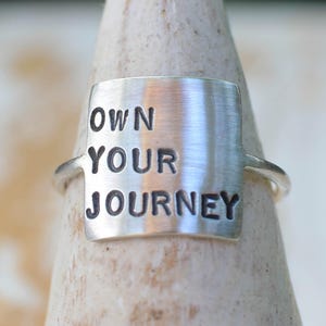 K&ouml;nnte beinhalten: Ein silberner Ring mit einer rechteckigen Silberplatte, auf der "OWN YOUR JOURNEY" in schwarzen Buchstaben steht.