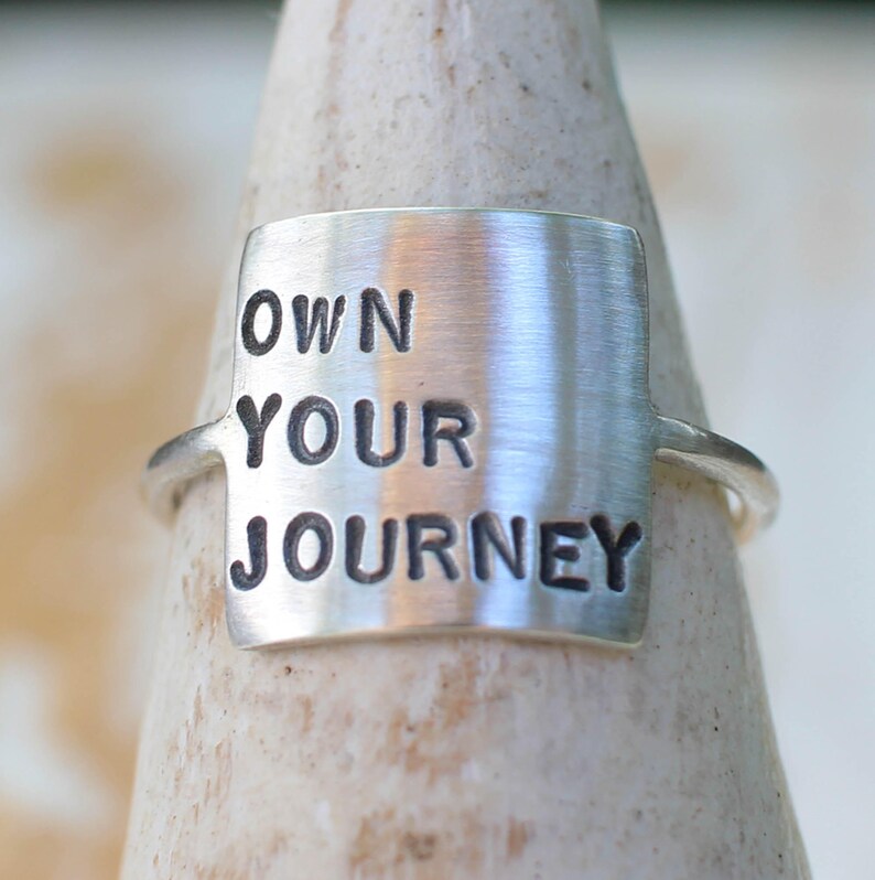 K&ouml;nnte beinhalten: Ein silberner Ring mit einer rechteckigen Silberplatte, auf der "OWN YOUR JOURNEY" in schwarzen Buchstaben steht.