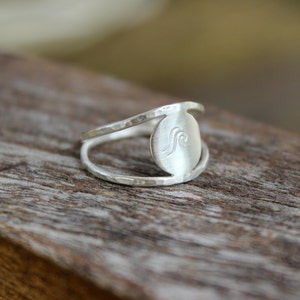 May include: A silver ring with a hammered finish and a circular design featuring a wave graphic.