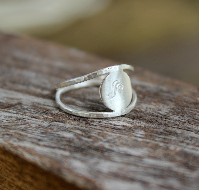May include: A silver ring with a hammered finish and a circular design featuring a wave graphic.