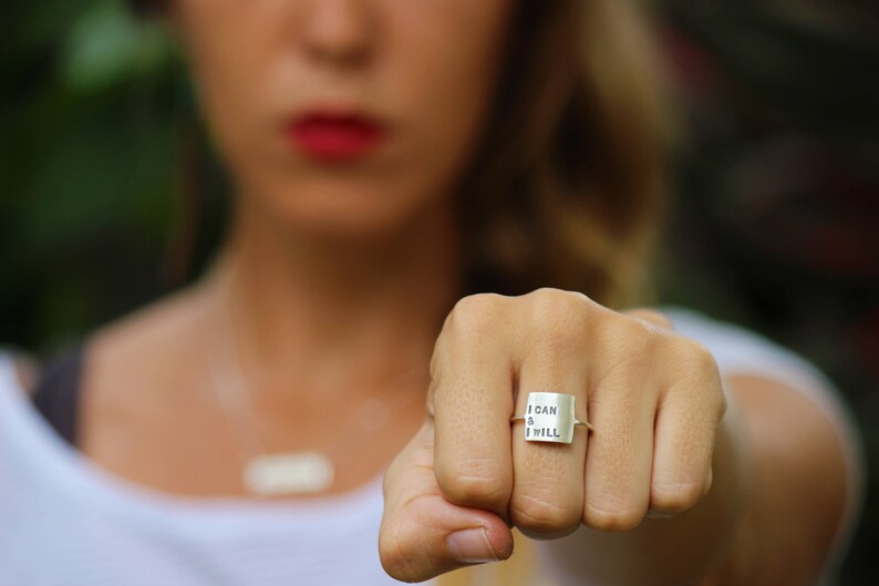 K&ouml;nnte beinhalten: Ein silberner Ring mit einem quadratischen Band, auf dem in einer einfachen Schrift "I CAN & I WILL" steht.