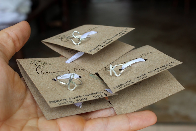 Op de afbeelding: Drie kaarten met een bruine papieren achtergrond en een wit lint. Elke kaart heeft een sterling zilveren initialenring met een andere letter. De tekst op de kaarten luidt "Sterling Silver Initial Ring Maat 8".