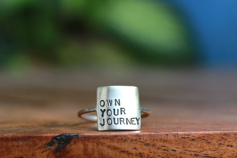 K&ouml;nnte beinhalten: Ein silberner Ring mit der Gravur "OWN YOUR JOURNEY" auf dem Band.