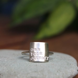 K&ouml;nnte beinhalten: Silberring mit einem quadratischen Band, auf dem "OWN YOUR JOURNEY" in erhabenen Buchstaben steht.