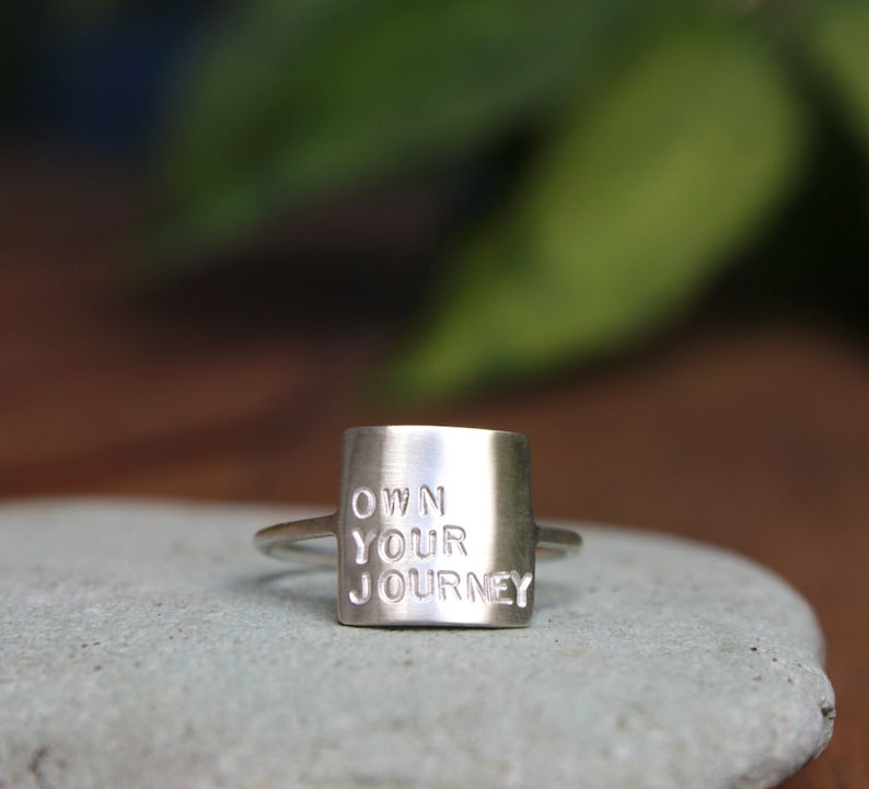 K&ouml;nnte beinhalten: Silberring mit einem quadratischen Band, auf dem "OWN YOUR JOURNEY" in erhabenen Buchstaben steht.