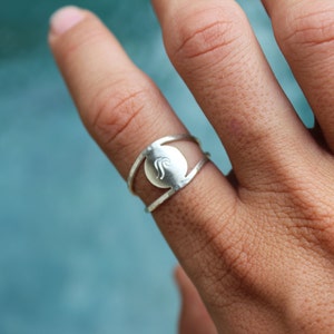 May include: A silver ring with a circular design featuring a wave-like pattern. The ring is worn on a finger.