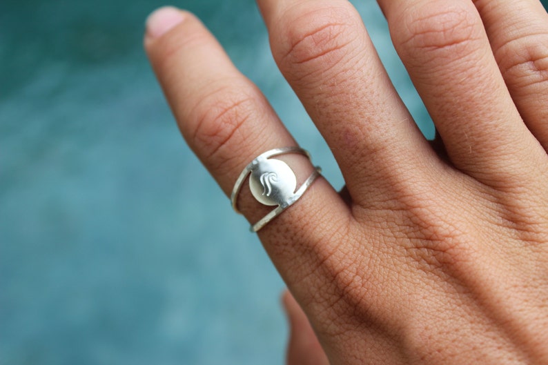 May include: A silver ring with a circular design featuring a wave-like pattern. The ring is worn on a finger.