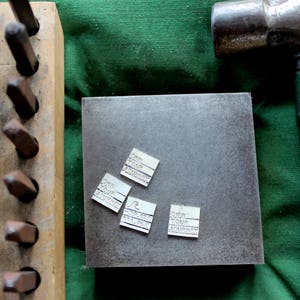 K&ouml;nnte beinhalten: Nahaufnahme einer Metalloberfl&auml;che mit f&uuml;nf kleinen silbernen Metalletiketten. Die Etiketten tragen den Text "Own Your Journey" aufgedruckt. Die Oberfl&auml;che liegt auf einem gr&uuml;nen Stoffhintergrund.