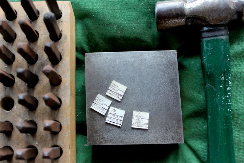 K&ouml;nnte beinhalten: Nahaufnahme einer Metalloberfl&auml;che mit f&uuml;nf kleinen silbernen Metalletiketten. Die Etiketten tragen den Text "Own Your Journey" aufgedruckt. Die Oberfl&auml;che liegt auf einem gr&uuml;nen Stoffhintergrund.
