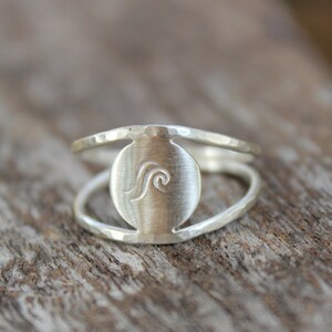 May include: A silver ring with a hammered finish and a circular cutout featuring a wave design.