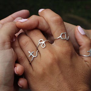 Op de afbeelding: Twee handen dragen zilveren ringen met verschillende letters en symbolen. De ringen zijn delicaat en hebben een gehamerde afwerking.