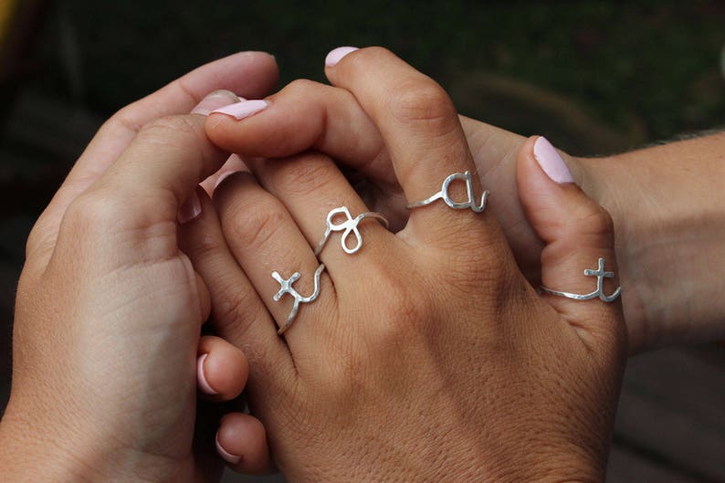 Op de afbeelding: Twee handen dragen zilveren ringen met verschillende letters en symbolen. De ringen zijn delicaat en hebben een gehamerde afwerking.