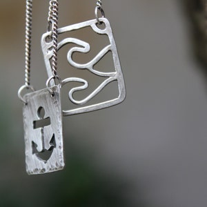 May include: Two silver pendant necklaces. One pendant features an anchor cutout, the other has a wave design. Both are rectangular and hang from delicate silver chains. The background is blurred, with a tree trunk visible.