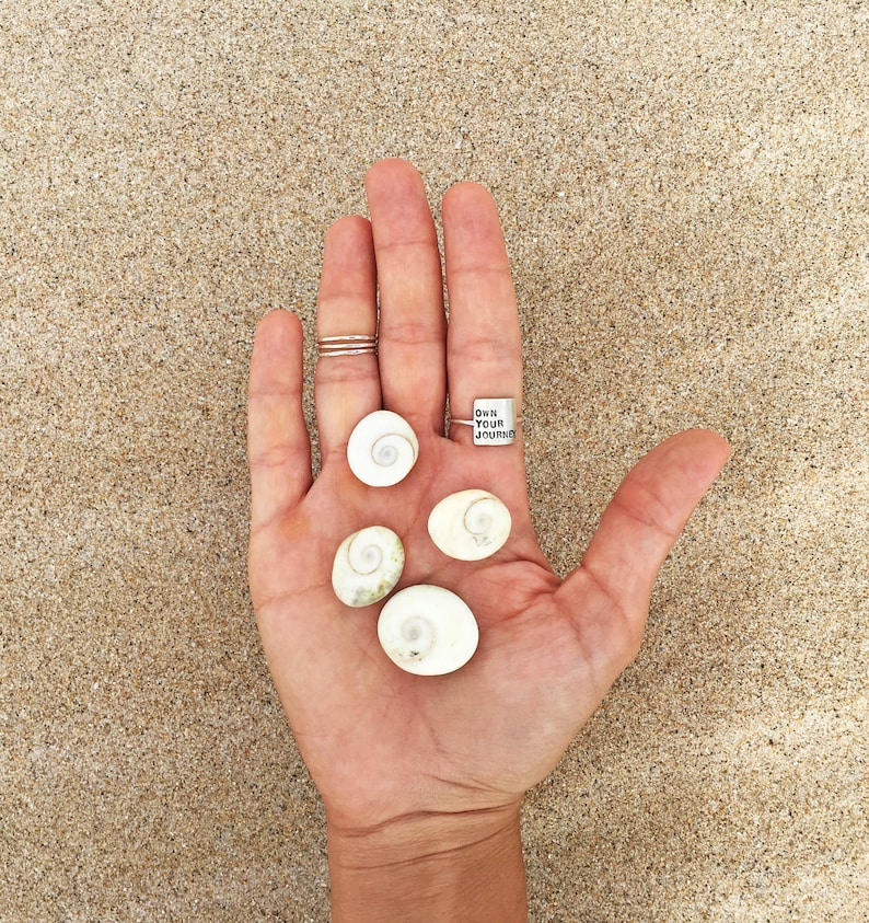 K&ouml;nnte beinhalten: Eine Hand h&auml;lt f&uuml;nf wei&szlig;e Muscheln und einen silbernen Ring mit der Aufschrift "Own Your Journey".
