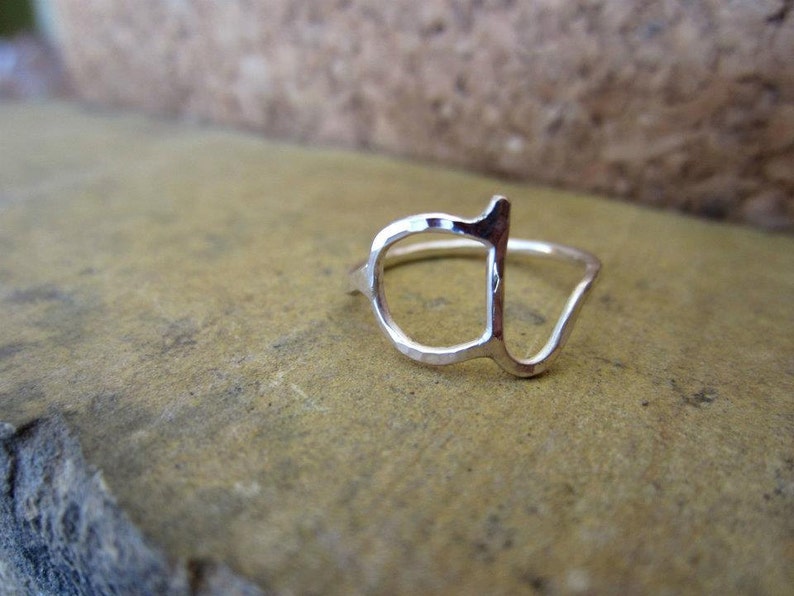Op de afbeelding: Een zilveren ring in de vorm van de letter 'a'. De ring is gehamerd en heeft een textuur.