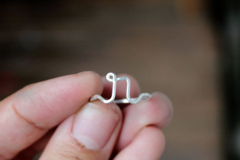 Op de afbeelding: Een zilveren ring met een eenvoudig, gebogen ontwerp. De ring is gemaakt van draad en is gevormd als de letter "J".