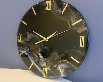 Peut inclure: Horloge en résine noire et blanche avec des chiffres romains dorés et des aiguilles dorées. Le cadran de l'horloge a un design marbré avec des tourbillons blancs et dorés.