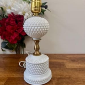 May include: A white hobnail table lamp with a brass base and accents. The lamp has a round, textured globe and a cylindrical base. The lamp is on a wooden surface, with a bouquet of red and white flowers in the background.