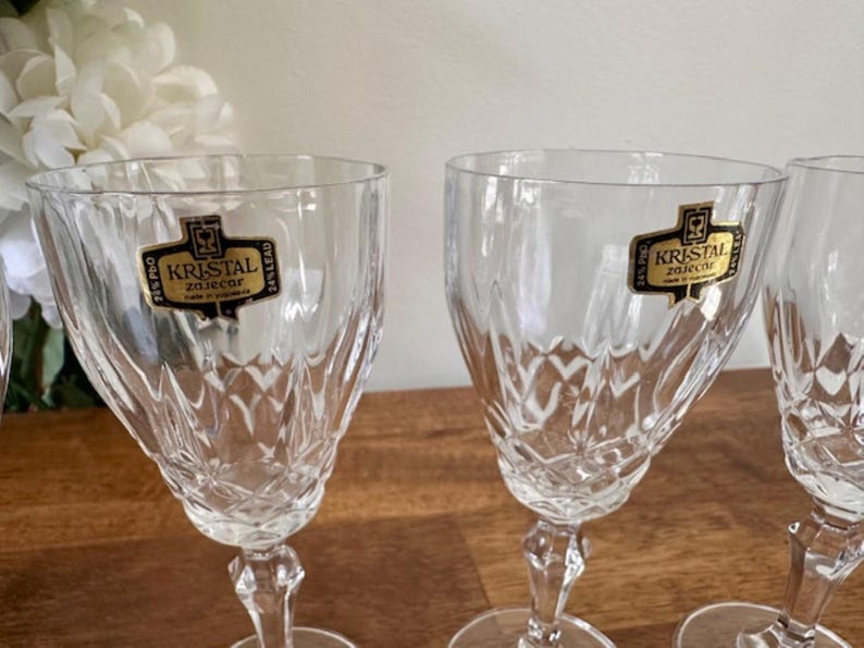May include: Four clear glass wine glasses with a cut crystal design. Each glass has a gold label that reads "Kristal Zaječar 24% Lead"