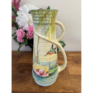 May include: Cream-coloured pitcher with a curved handle and a detailed painted design. The design features a kingfisher bird, water lilies, and reeds, with a green and yellow landscape. The pitcher has a textured surface and a green rim.