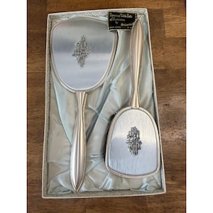 May include: A vintage dressing table set by Masco, featuring a hand mirror and a brush. Both items have silver-toned metal frames and handles, with an ornate, jewel-like embellishment on the mirror and brush. The set is presented in a box with a satin lining.