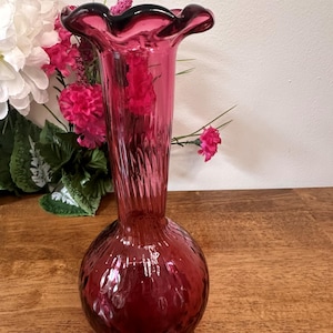 Peut inclure: Un vase en verre rouge rubis avec un bord festonné et une base bulbeuse. Le vase présente des détails côtelés verticaux sur le col et le corps. Il est posé sur une surface en bois, avec des fleurs roses en arrière-plan.