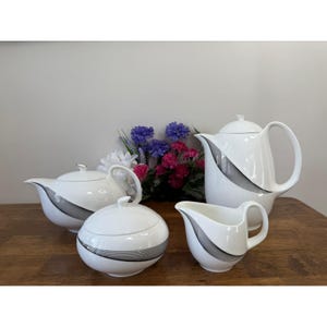 Peut inclure: Service à thé en porcelaine blanche avec un motif de rayures noires et grises. L'ensemble comprend une théière, une crémière, un sucrier et une deuxième théière. L'ensemble est présenté sur une surface en bois avec des fleurs en arrière-plan.