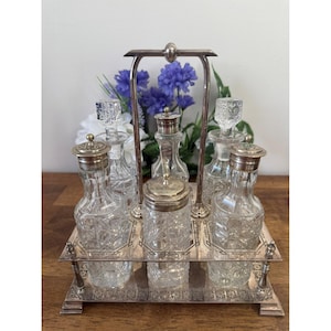 May include: A silver-toned condiment set with six clear glass bottles and jars. The bottles have decorative stoppers and are arranged on a silver tray with a handle. The set is likely vintage and suitable for serving condiments.