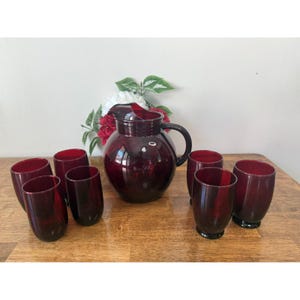 Peut inclure: Un pichet en verre rouge rubis vintage avec une poignée et un ensemble de gobelets assortis. Le pichet et les verres sont disposés sur une surface en bois. La verrerie est d'un rouge profond et transparent.