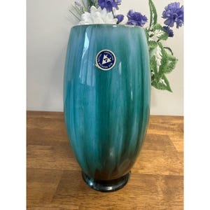 Puede incluir: Un jarrón alto de color verde azulado con un acabado brillante. El jarrón tiene una base oscura y una etiqueta que dice "Blue Mountain Pottery". El jarrón está lleno de flores artificiales blancas y moradas. El jarrón mide unos 25 cm de alto.