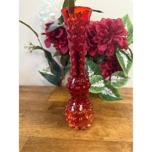 May include: A tall, red glass vase with a textured, hobnail design. The vase has a flared top and a rounded base. The background features red and white flowers with green foliage, set on a wooden surface.