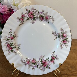 Puede incluir: Plato de porcelana blanca con borde festoneado y ribete dorado. El plato está decorado con rosas rosas y hojas verdes. El plato se exhibe sobre un soporte dorado. Un ramo de flores rosas y blancas está en el fondo.