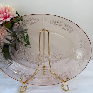 May include: A pink oval serving platter with floral detailing, displayed on a gold-coloured metal stand. The platter has an embossed floral pattern around the rim and centre. The stand has a curved design.