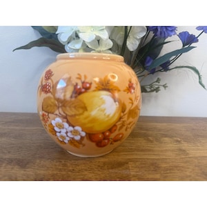 May include: A peach-colored, round ceramic vase with a fruit design. The vase features illustrations of yellow apples, red berries, and white flowers. The vase is sitting on a wooden surface.